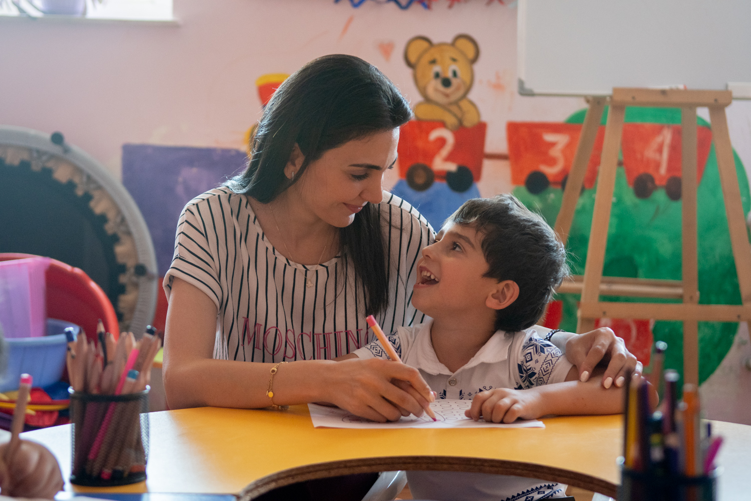 Early education center. Shriven. The caregiver is helping a boy while he is doing a drawing. The strong emotional connection between the caregiver and the patience has a strong impact in the benefits of the practice.